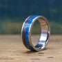 A titanium ring featuring an inlay of textured dark meteorite and vibrant blue opal stands upright on a rustic wooden surface.