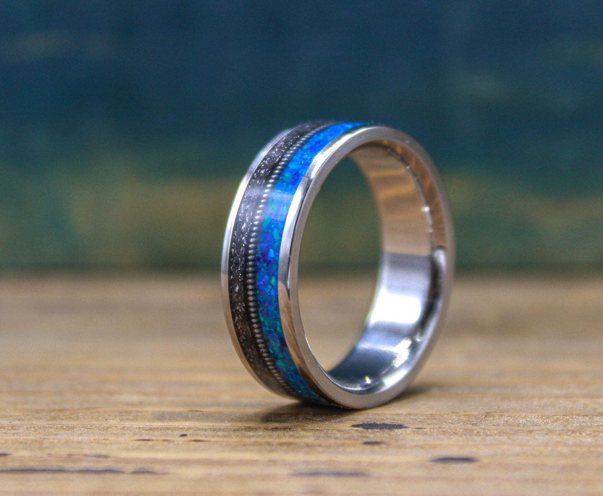 A titanium ring featuring an inlay of textured dark meteorite and vibrant blue opal stands upright on a rustic wooden surface.