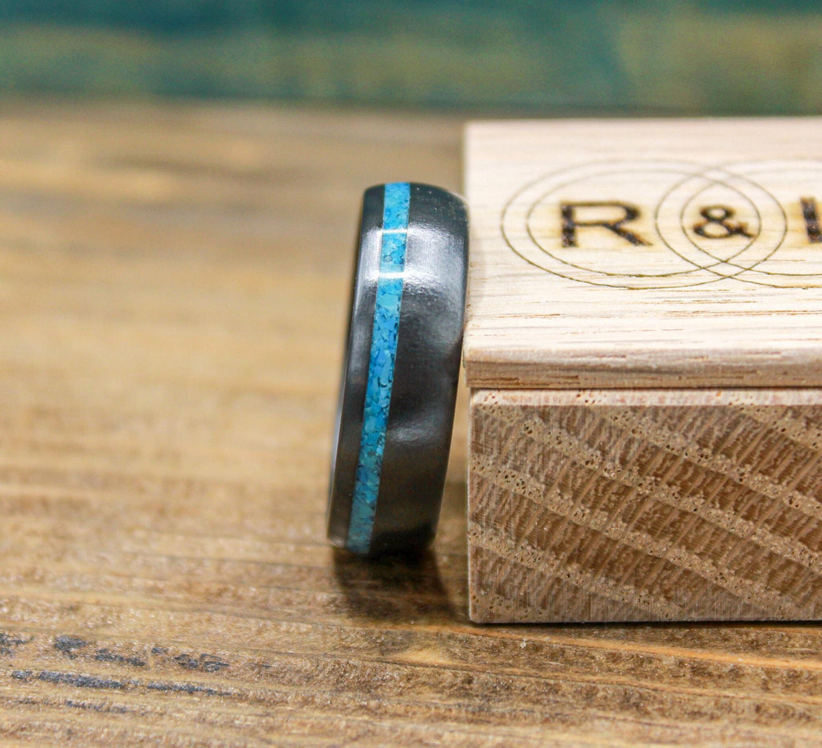 A dark ring with a turquoise inlay stands next to a wooden box with a circular monogram.