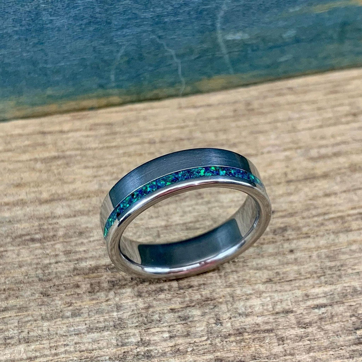 A brushed titanium band featuring a vibrant green and blue opal inlay sitting on a rustic wooden board.