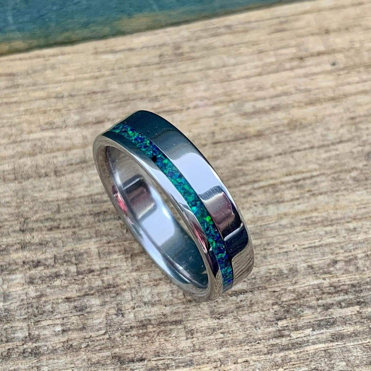 An angled view of a polished metal ring with a central green and blue opal stripe on a wooden background.