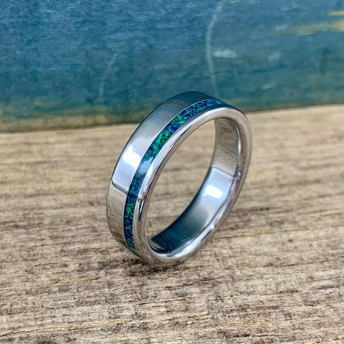 A close-up shot of a polished band with a thin blue-green opal inlay resting vertically on wood.