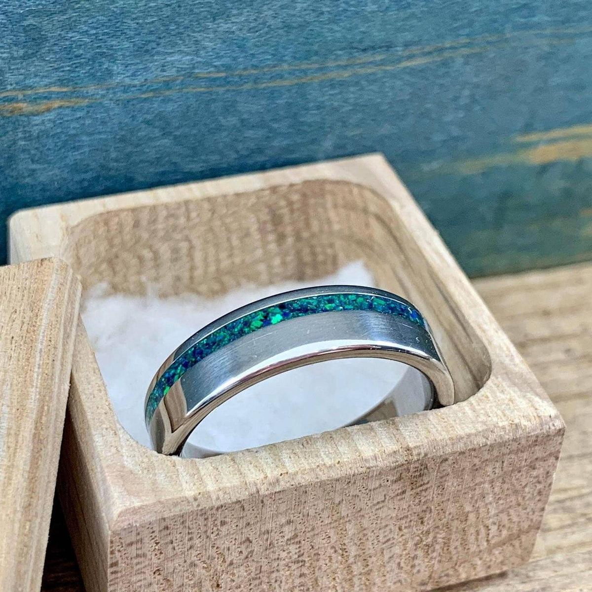 A close-up of a brushed titanium ring with a green-blue opal inlay placed inside a small wooden box.
