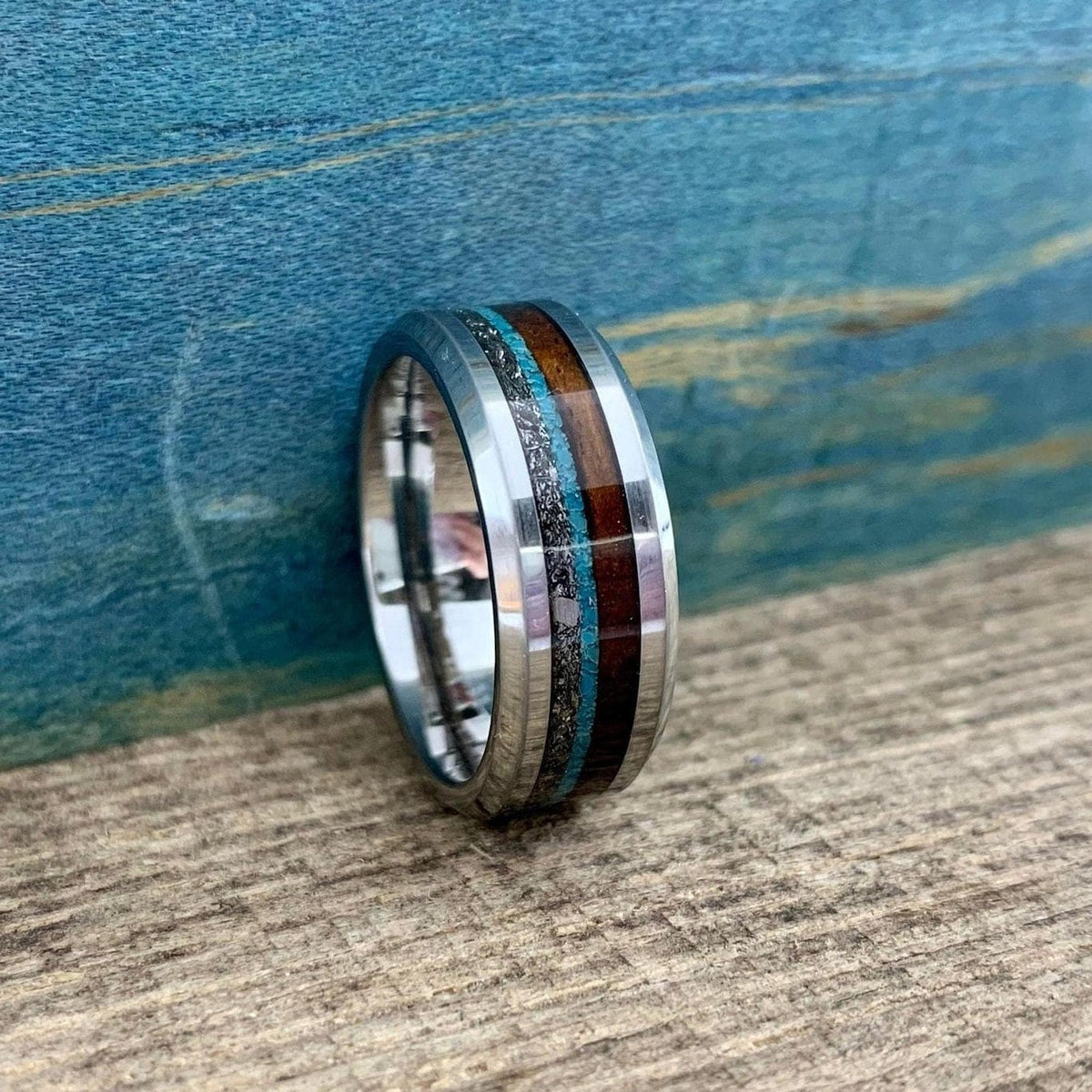 A metal ring featuring inlays of wood, meteorite, and turquoise resting on a wooden surface against a blue backdrop.