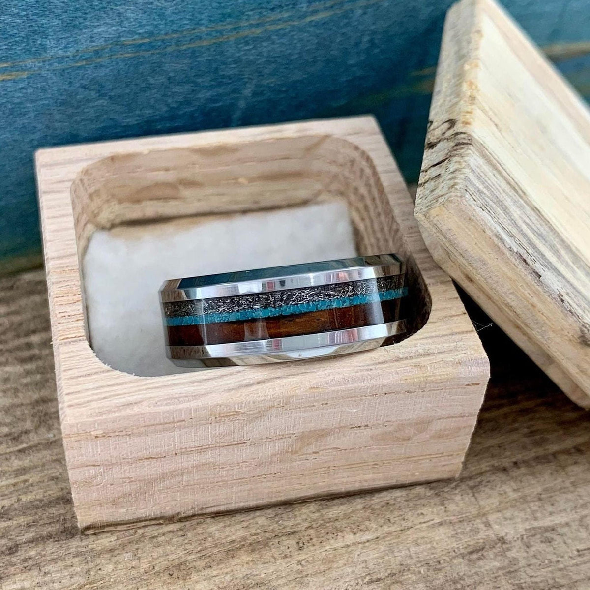 A metal ring with wood and turquoise inlays resting inside an open light-colored wooden box.