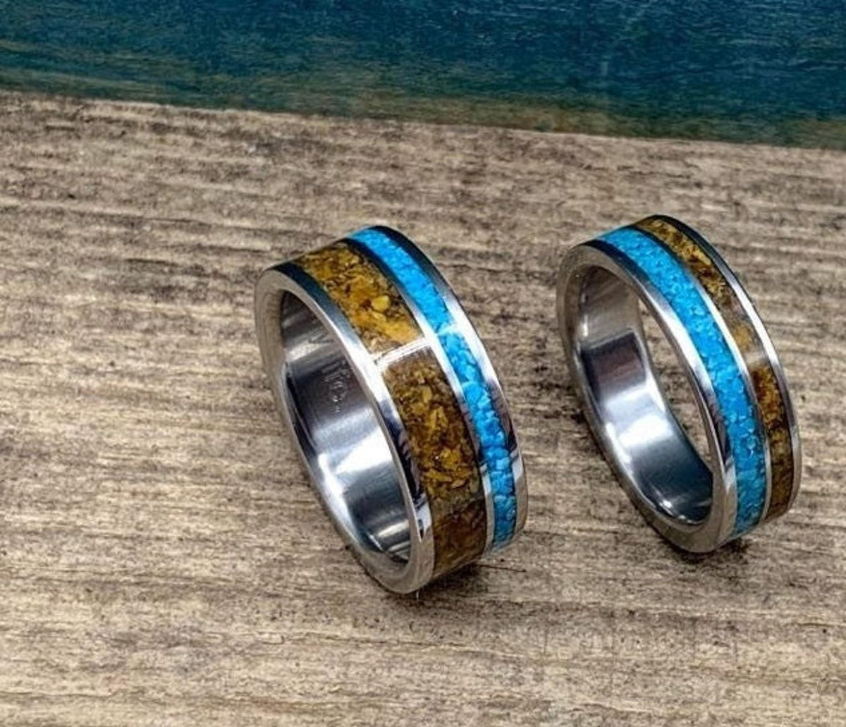 Two metal rings with turquoise and tiger's eye inlays placed side by side on a wooden tabletop.