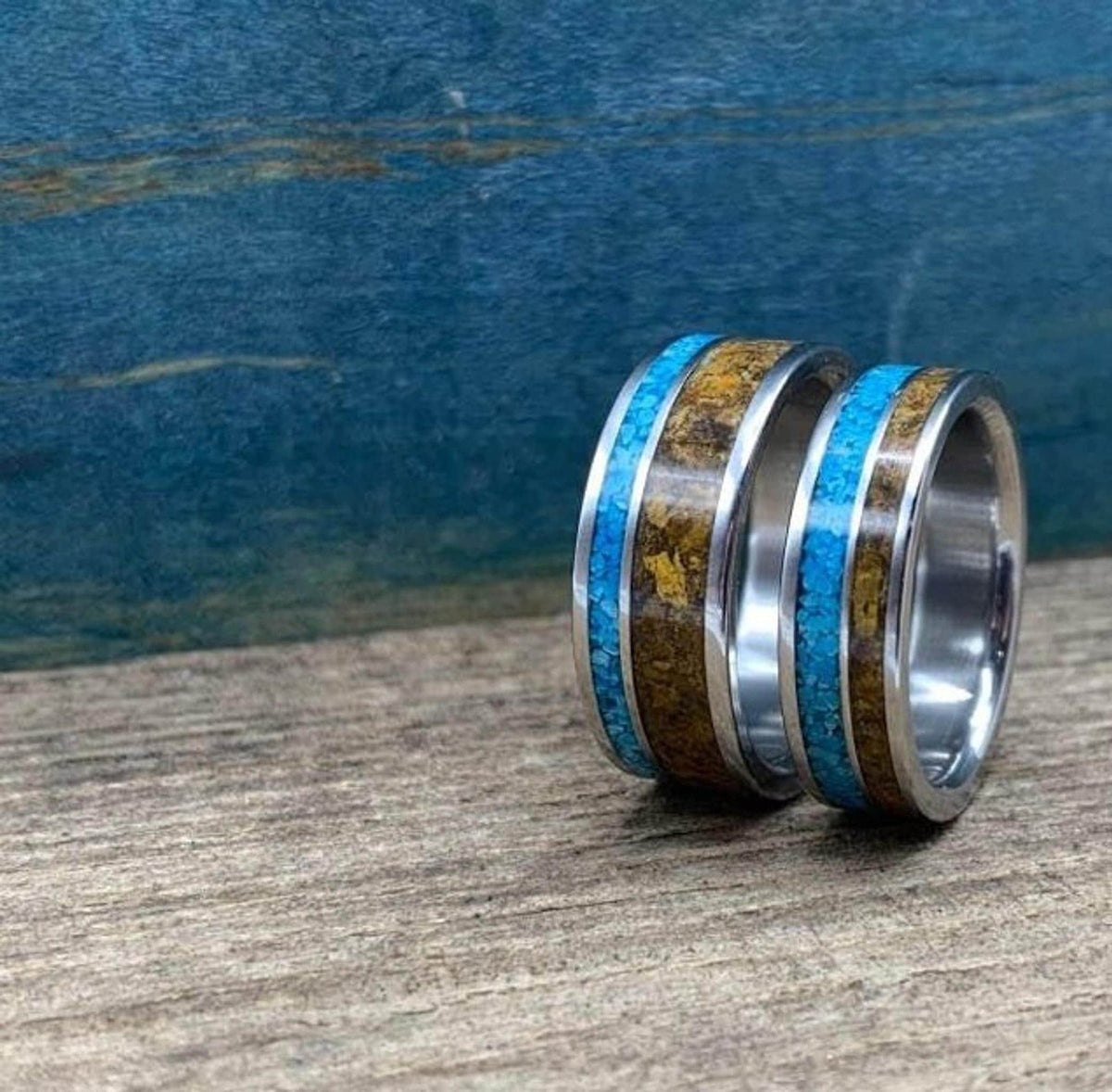 Two metal rings featuring crushed turquoise and tiger's eye inlays, standing upright against a blue background.