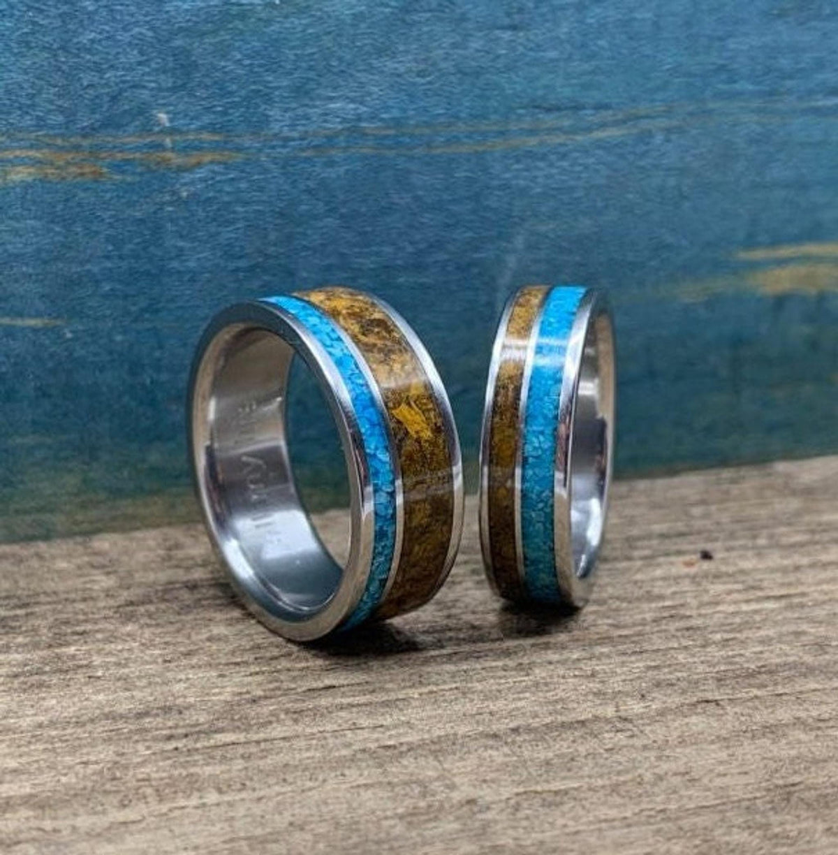 Two matching metal rings inlaid with turquoise and brown tiger's eye, positioned upright on a rustic wooden board.