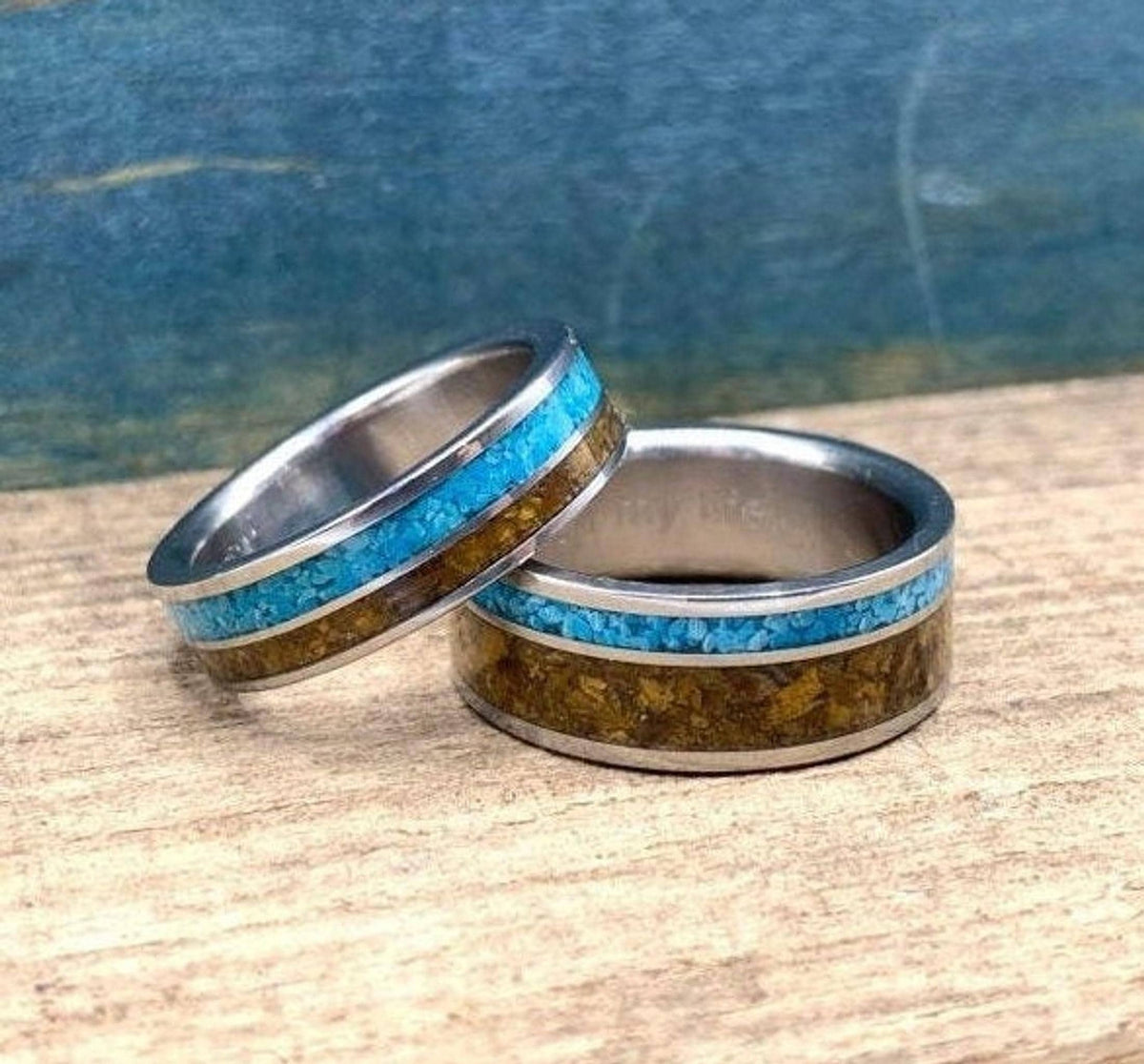 Two metal rings with inlaid turquoise and tiger's eye, with one ring slightly overlapping the other on a wooden background.