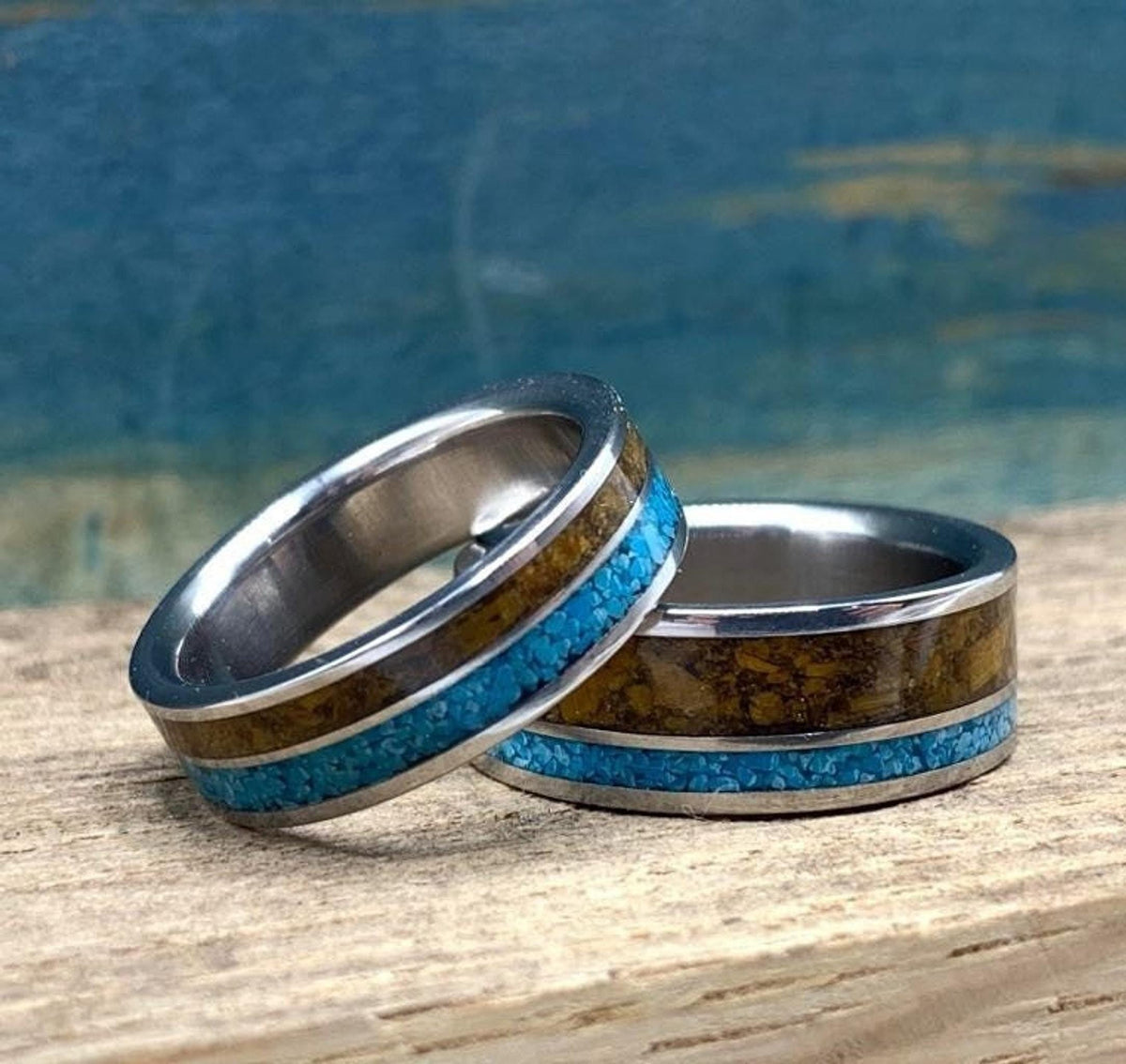 Two metal rings featuring turquoise and tiger's eye inlays, positioned side by side on a weathered wooden surface.