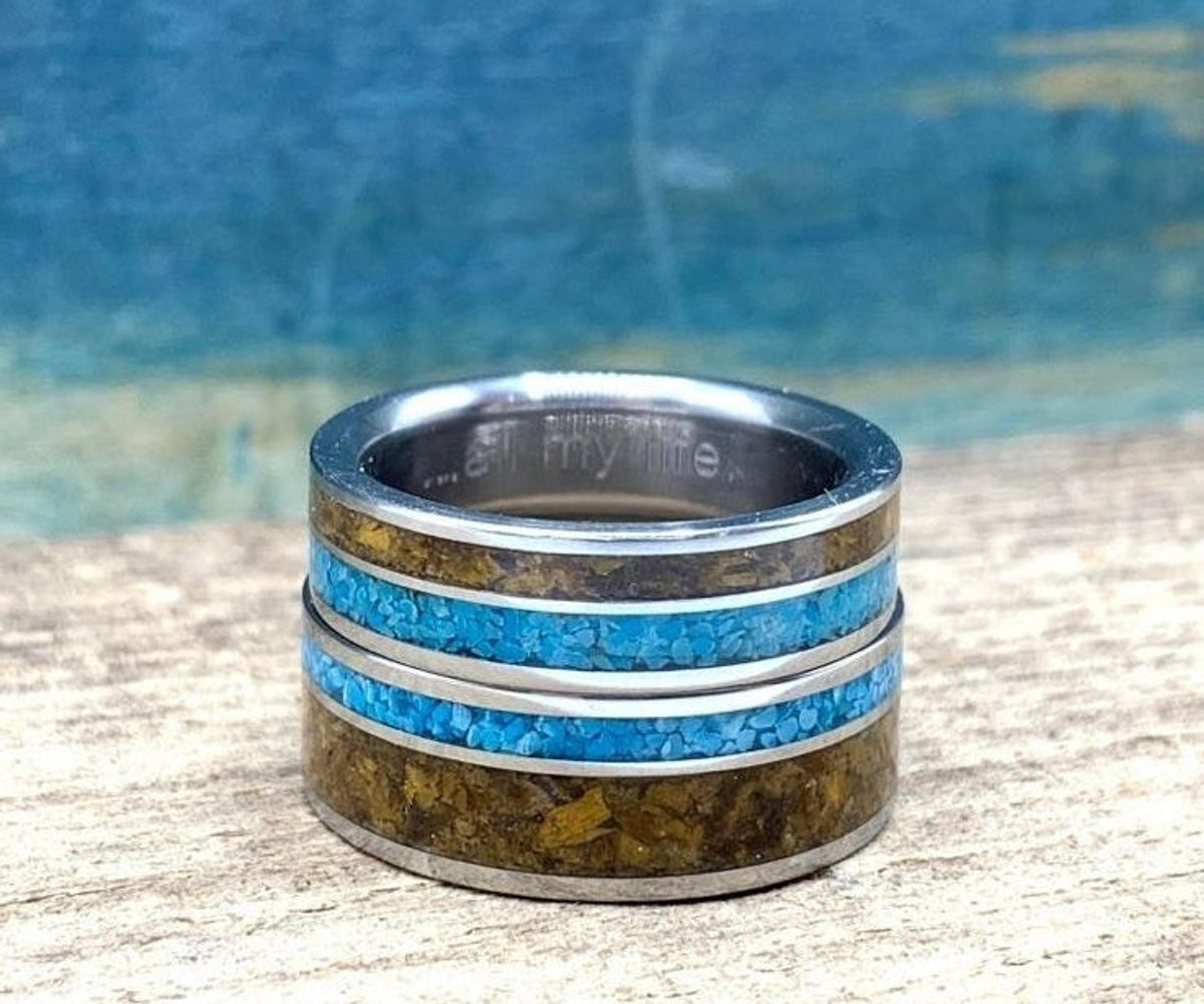 A single wide metal ring featuring two rows of turquoise inlay and two rows of tiger's eye inlay, showing faint engraved text on the inner band.