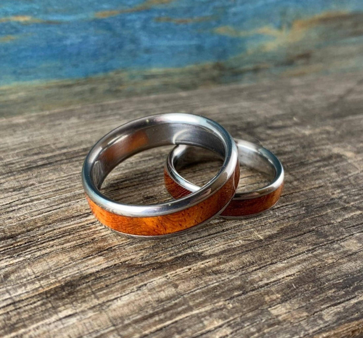 Two metal wedding bands featuring wood grain inlays lie nested together on a wooden tabletop.