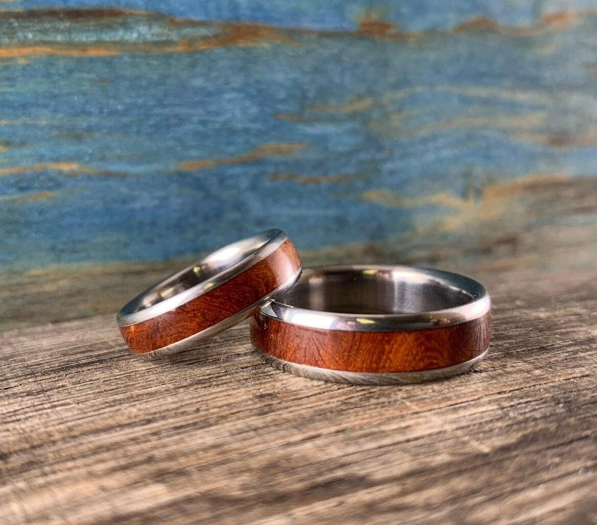 Two narrow and wide metal bands with reddish wood inlays sit side by side on weathered wood.
