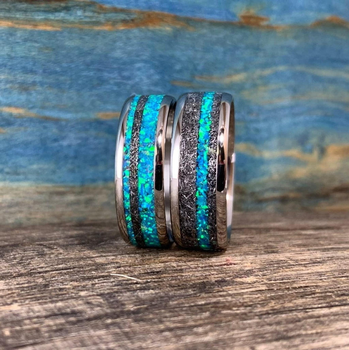 Two titanium wedding bands with alternating meteorite and bright blue-green opal inlay patterns stand upright on a wooden surface.