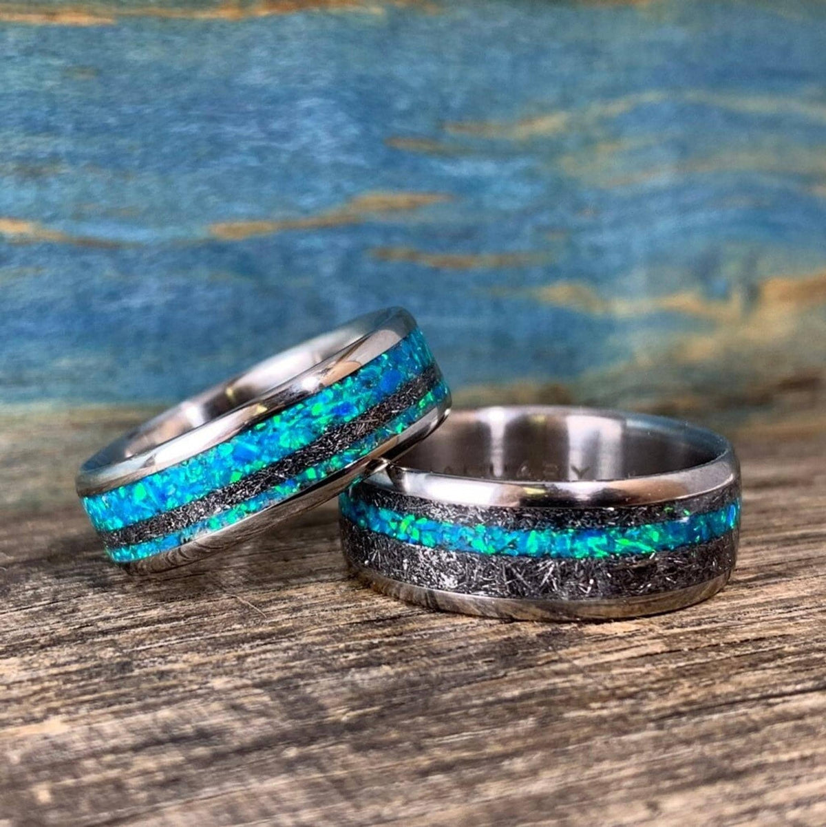 Two titanium wedding bands with meteorite and blue-green opal inlay sit side by side on a rustic wooden texture.