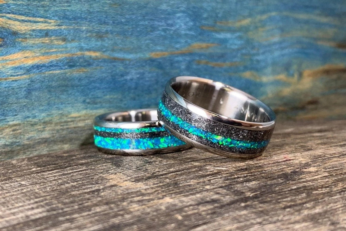 Two titanium rings featuring meteorite texture and vibrant blue-green opal stripes resting on a textured wooden surface.