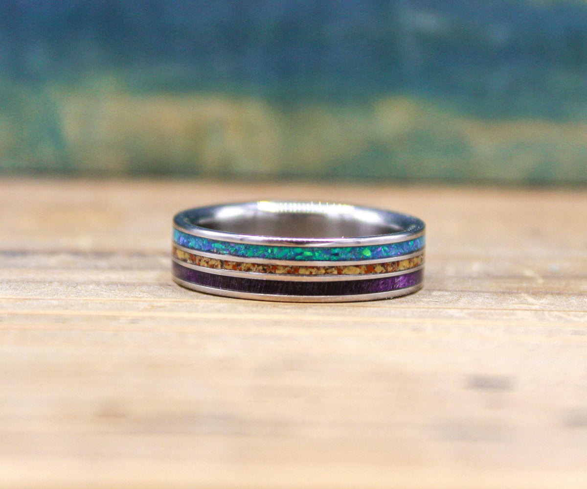 A titanium ring featuring three distinct inlays of blue opal, speckled dinosaur bone, and purple wood, resting on a wooden surface.