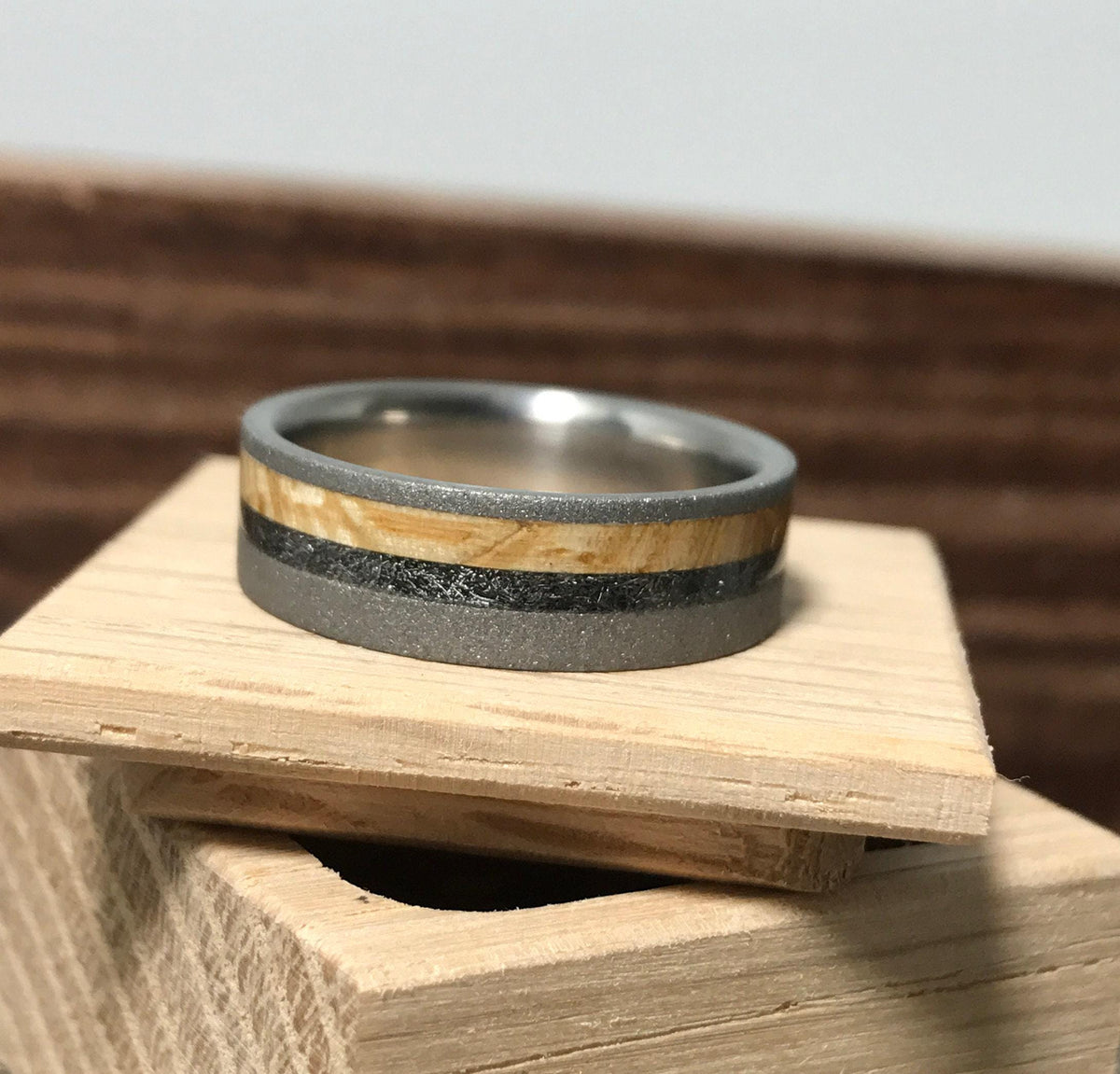 A sandblasted titanium wedding band featuring wood and meteorite inlays rests on a small wooden block.