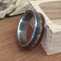 A custom ring featuring inlays of meteorite, dinosaur bone, and ironwood, standing upright on a wooden surface next to a small wooden block.