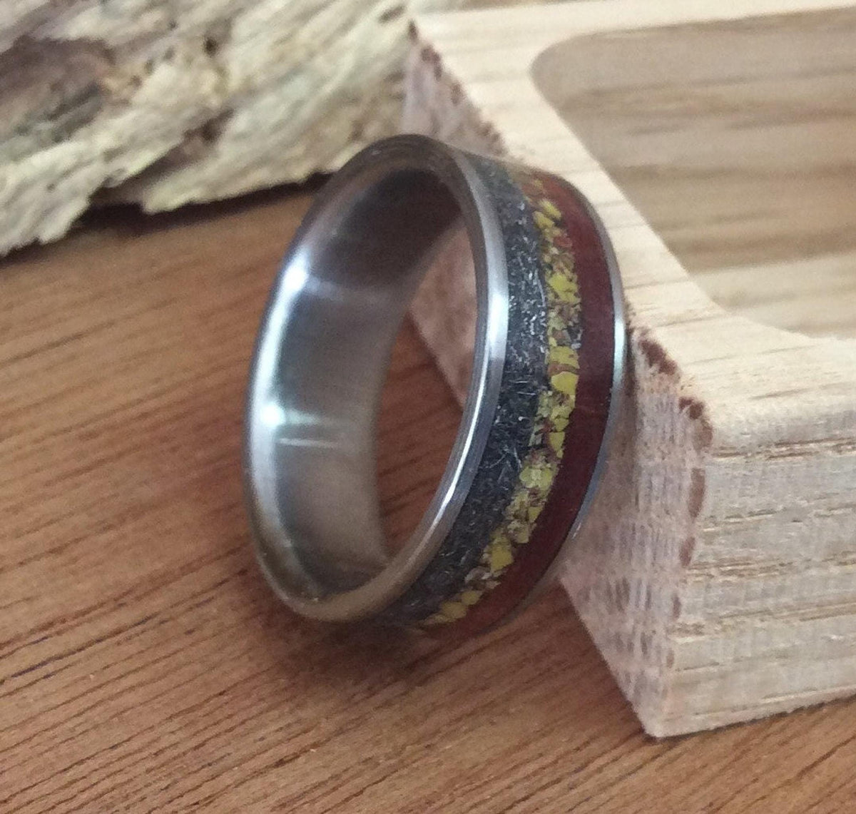A custom ring featuring inlays of meteorite, dinosaur bone, and ironwood, standing upright on a wooden surface next to a small wooden block.