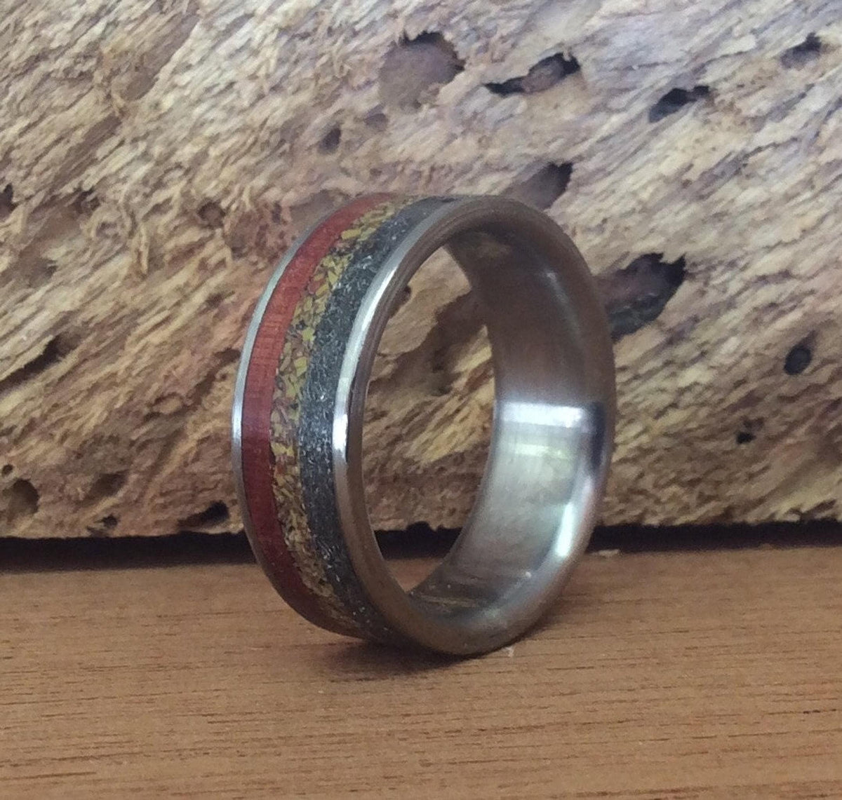 A band ring with dark metal edges and three central inlay bands of red wood, speckled dinosaur bone, and textured meteorite, positioned in front of a textured piece of wood.