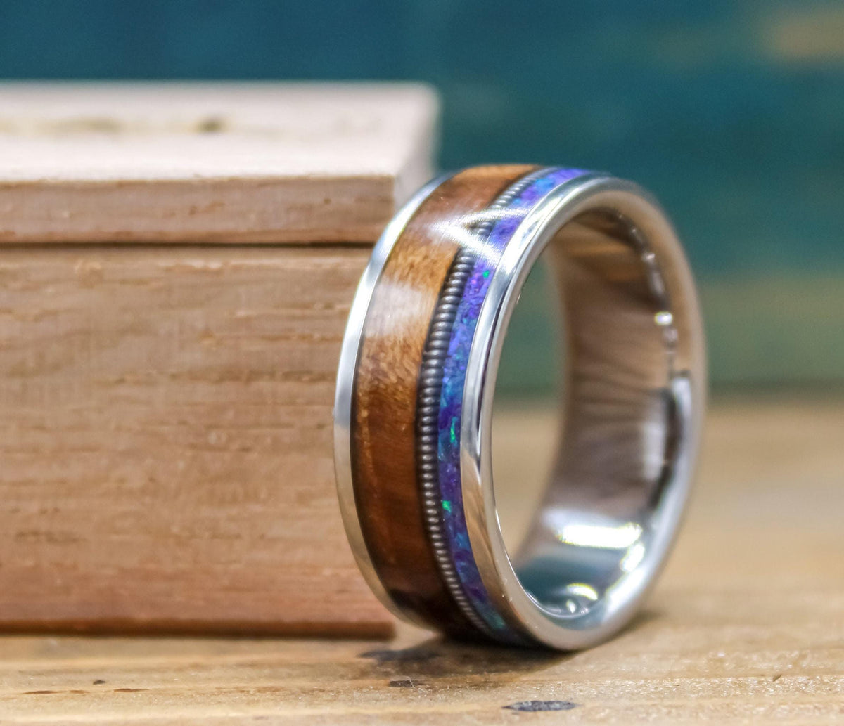 A titanium ring with wood and purple-blue opal inlay stands upright next to a wooden box.