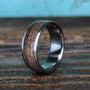 A close-up shot of a men's ring featuring a dark wood inlay bordered by silver-colored metal, resting on a textured wooden surface against a blue-and-brown backdrop.