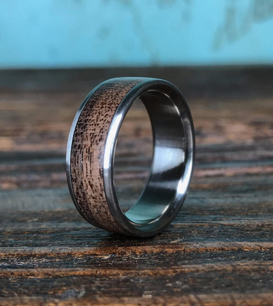 A close-up shot of a men's ring featuring a dark wood inlay bordered by silver-colored metal, resting on a textured wooden surface against a blue-and-brown backdrop.