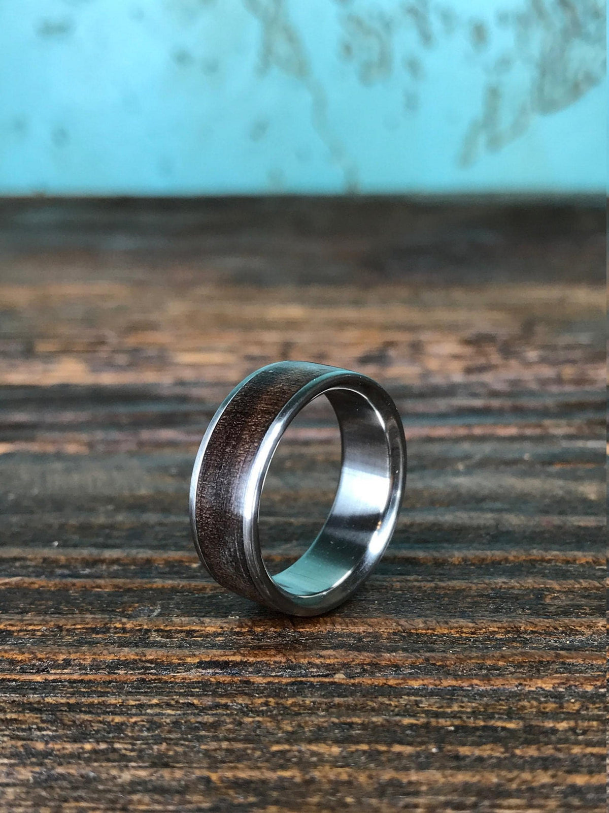 A ring with a dark wood inlay and silver-toned edges stands upright on a weathered wooden plank in front of a mottled blue and brown background.