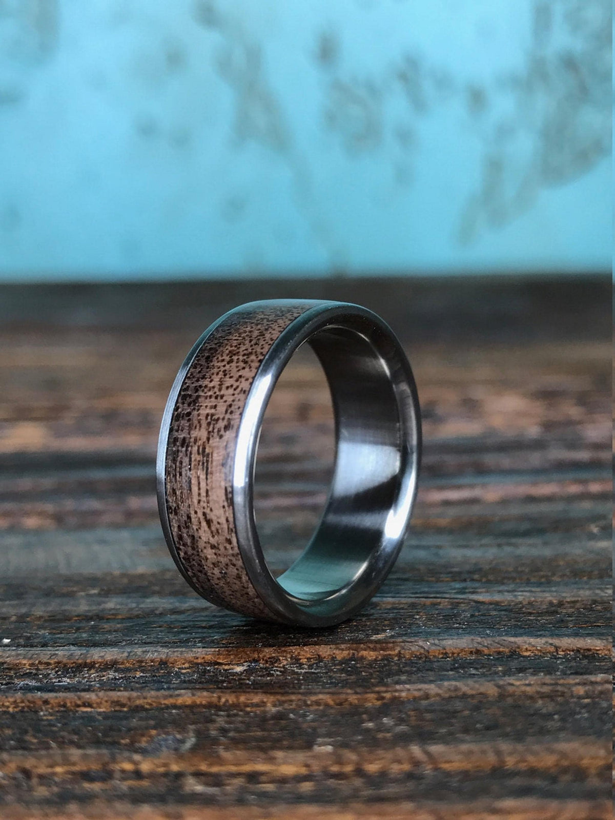 A ring with a textured dark wood inlay and metallic edging is positioned on a dark wooden surface with a soft blue backdrop.