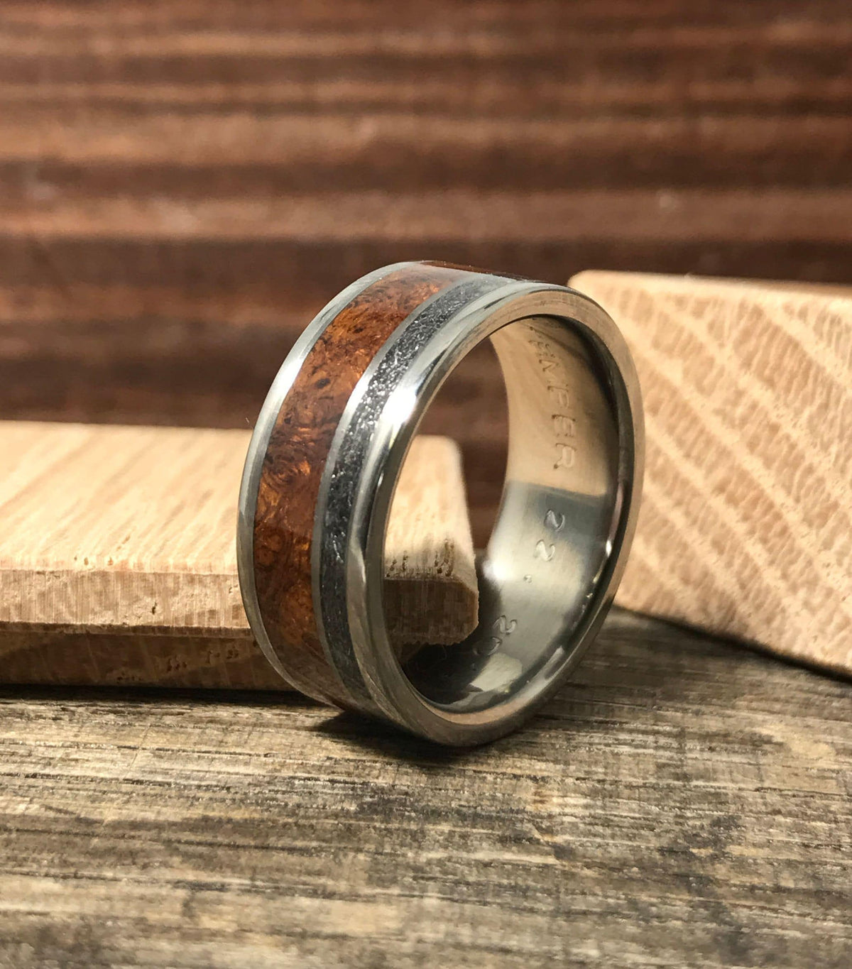 A titanium ring with wood and meteorite inlays is displayed between two wooden blocks, showing engraved text inside.