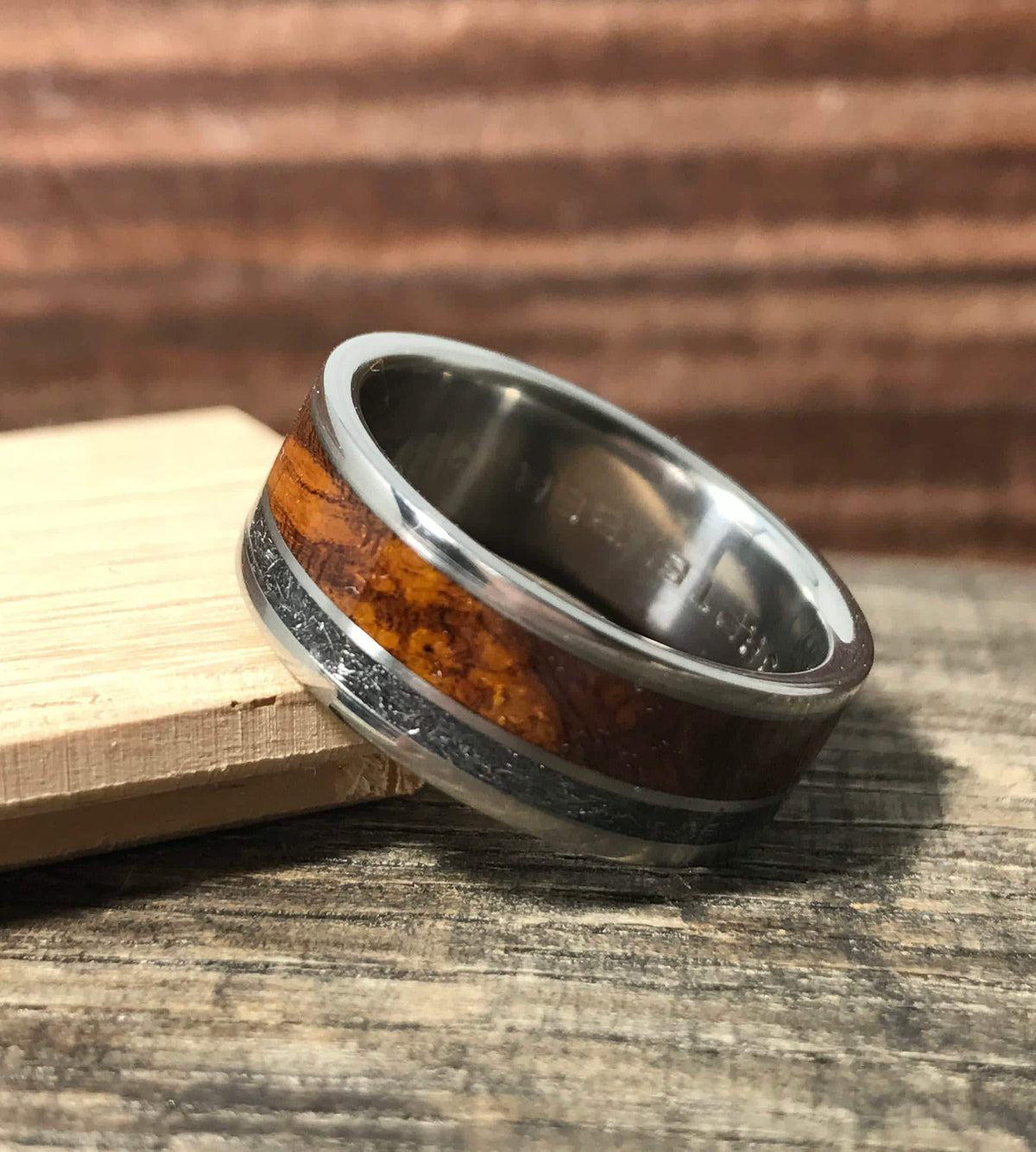 A titanium ring featuring a wood grain inlay and a textured meteorite stripe leans against a wooden block.