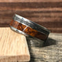 A titanium ring featuring an inlaid band of textured meteorite and rich brown wood resting on a light wood block.