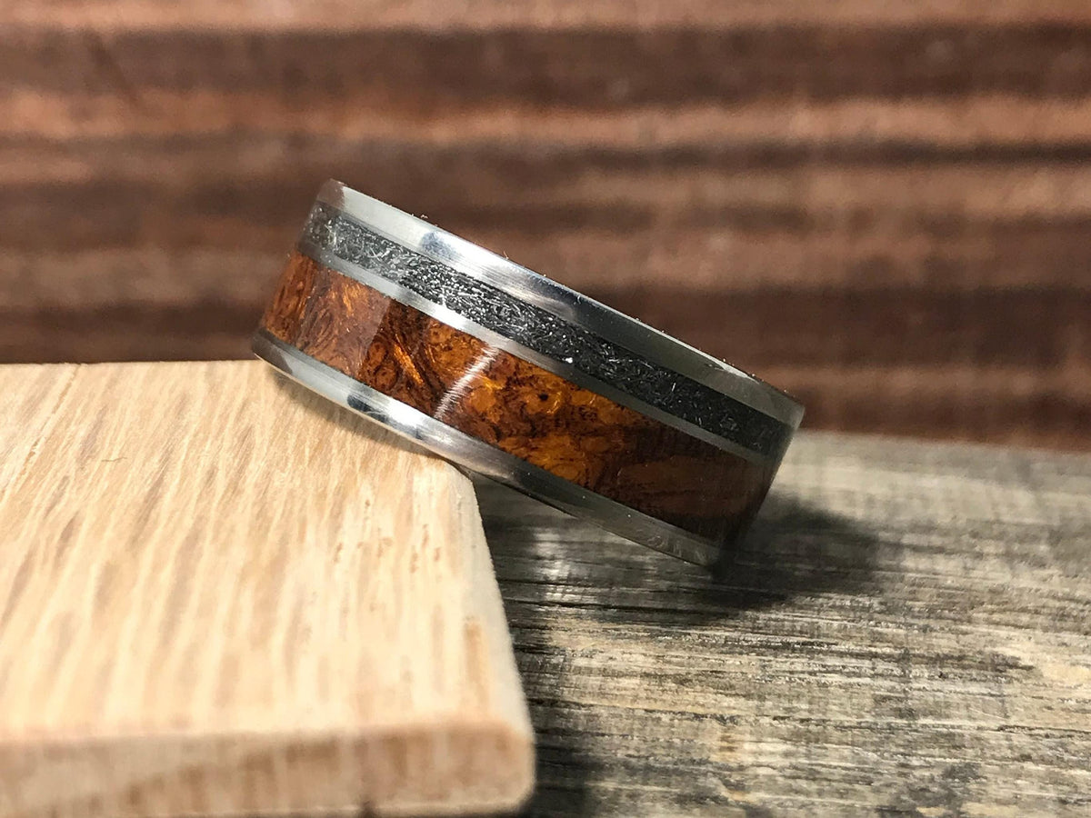 A titanium ring featuring an inlaid band of textured meteorite and rich brown wood resting on a light wood block.