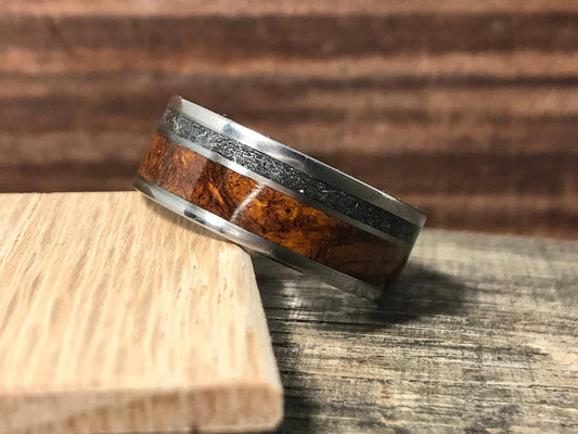 A titanium ring featuring an inlaid band of textured meteorite and rich brown wood resting on a light wood block.