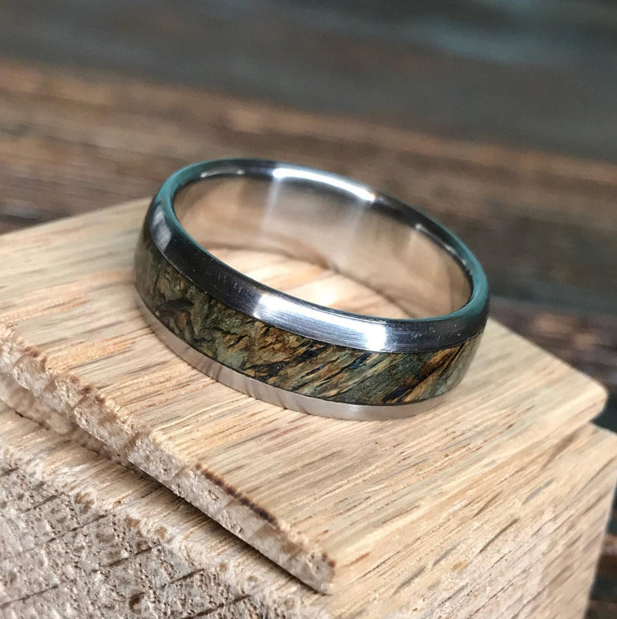 A titanium ring with a black ash burl wood inlay rests on a small piece of light-colored wood.