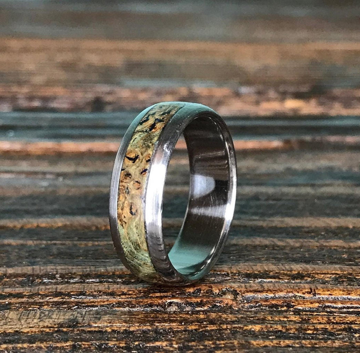 A titanium ring with a black ash burl inlay stands upright on a dark wooden surface.