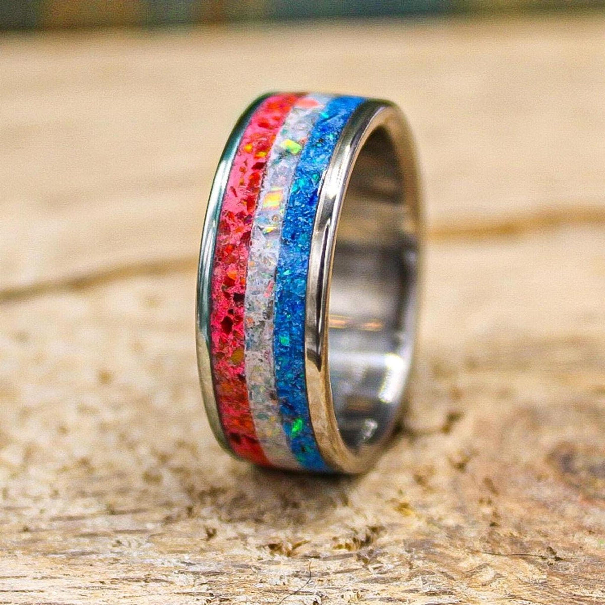 A custom titanium ring resting upright on a rustic wooden surface, featuring a three-stripe inlay of red, white, and blue opal material.