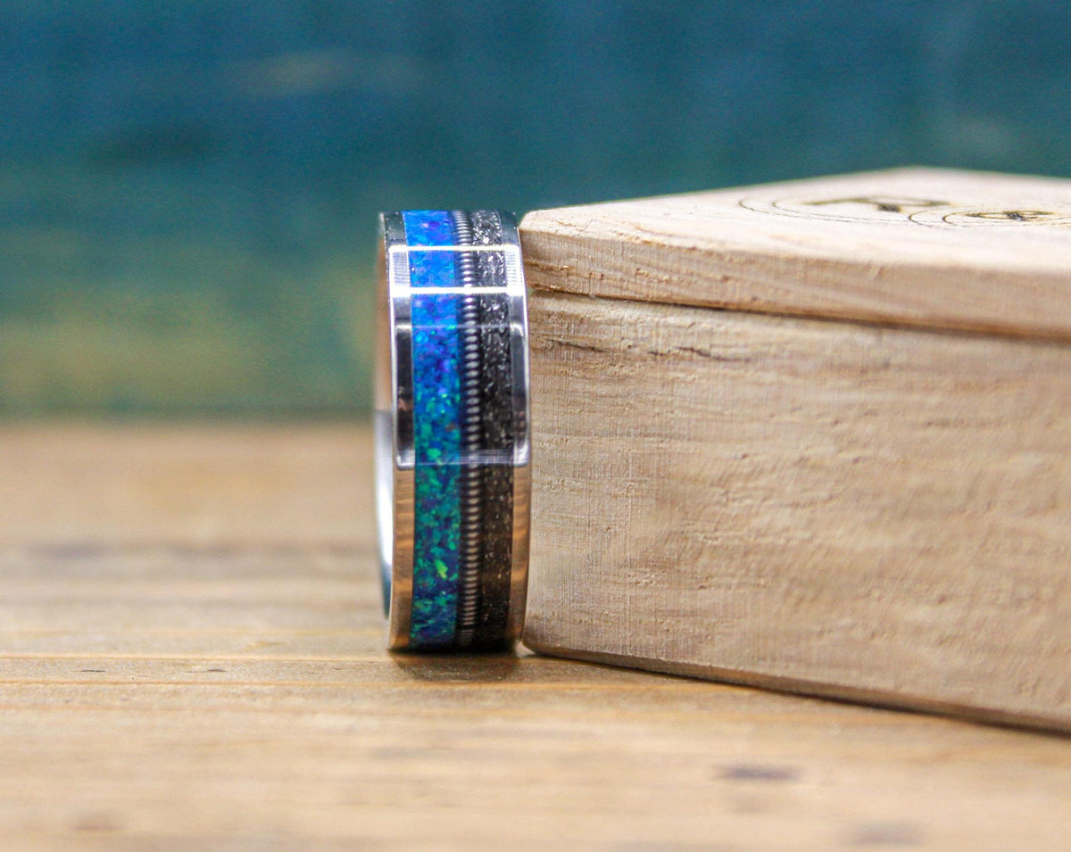 The titanium ring stands next to a small wooden box, displaying its vibrant blue opal and textured meteorite inlays.