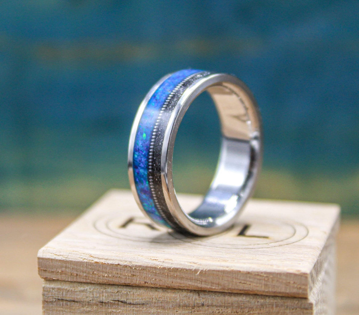 The ring stands on a branded wooden box, angled to highlight the bright blue opal inlay next to a thin metallic strip.