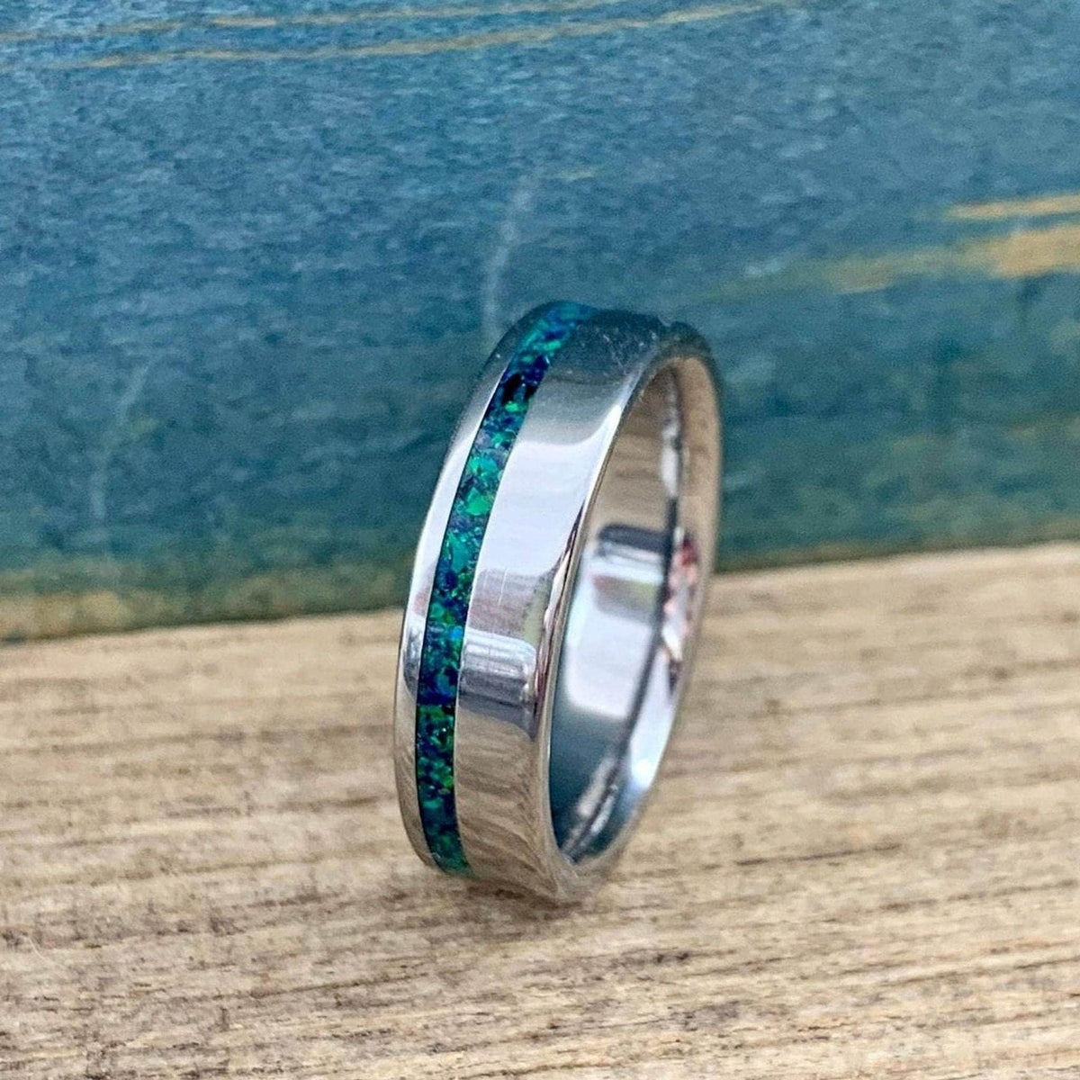 A polished titanium ring with a green and blue opal inlay rests on a wooden surface against a blue textured background.