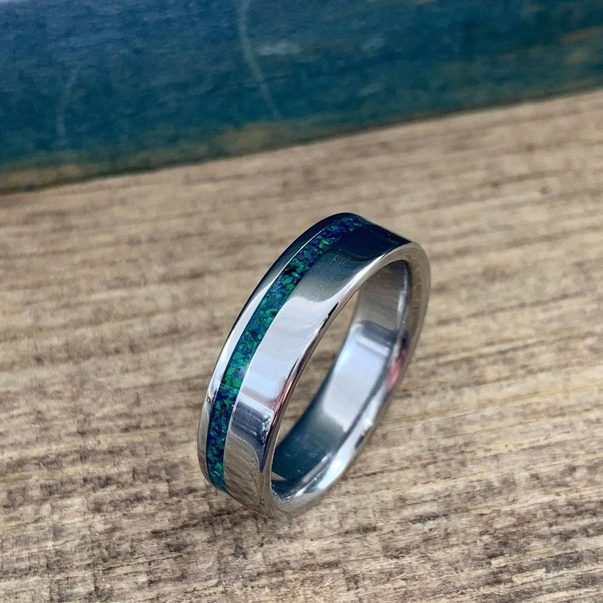 A polished metal ring displaying a shimmering green and blue opal inlay on a rustic wooden surface.