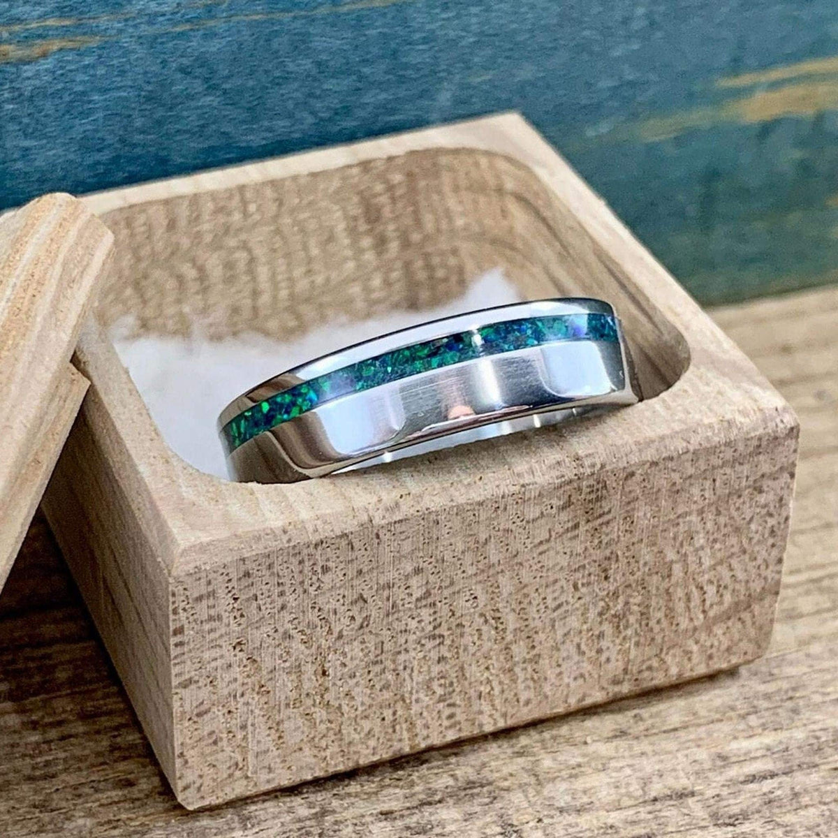 A metallic ring with a green and blue opal inlay resting inside an open wooden gift box.