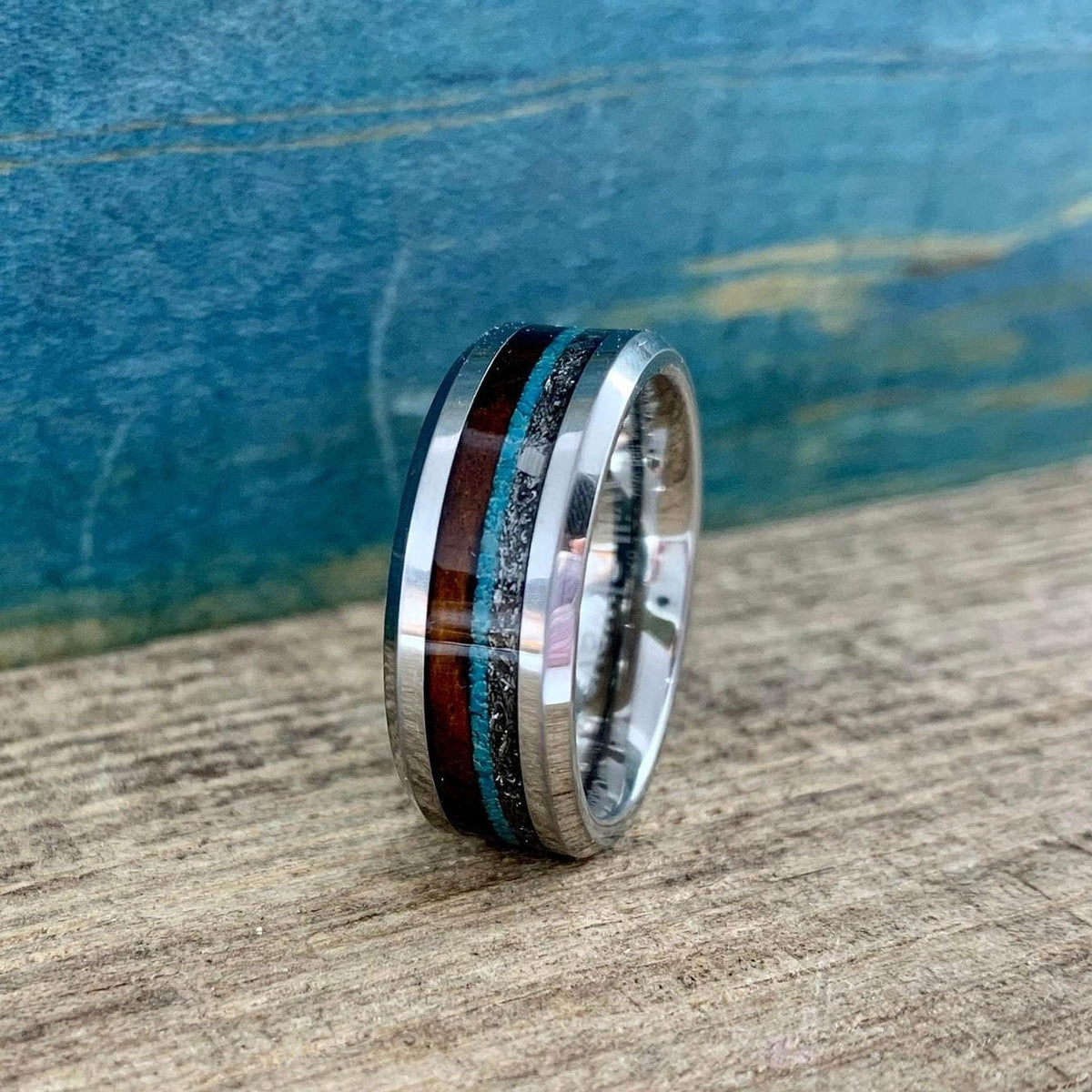 A metal ring with wood, meteorite, and turquoise inlays angled slightly left on a rustic wooden surface.