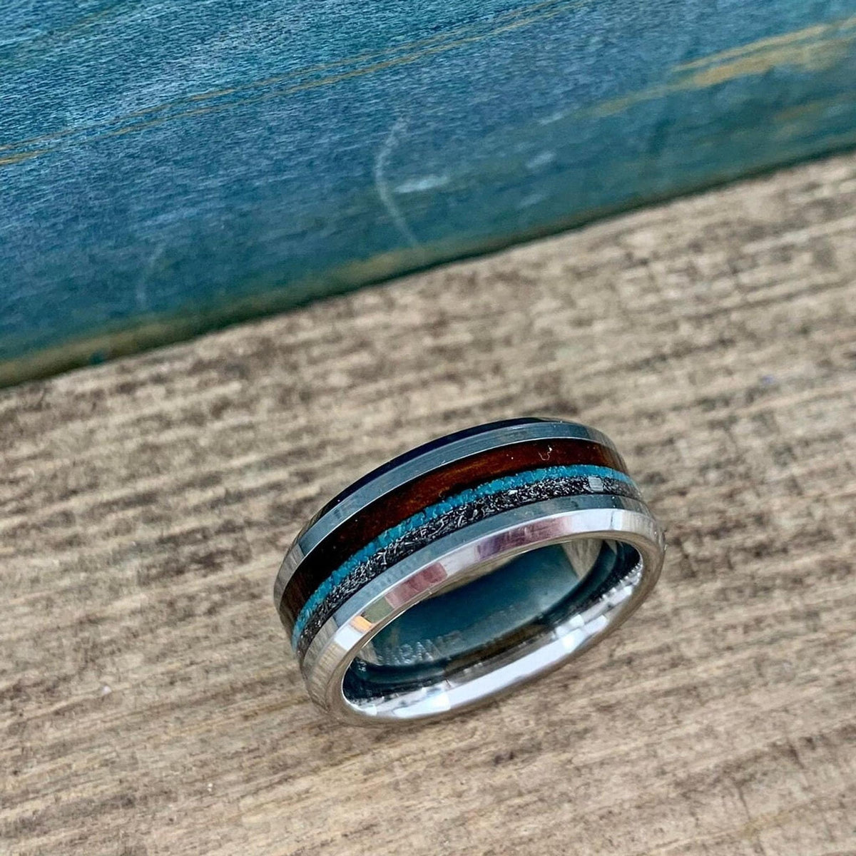 A banded ring with wood, metallic, and blue stone inlays lying at an angle on a textured wooden board.