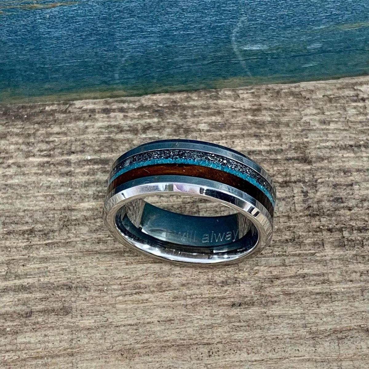 A metal ring with inlaid materials displayed horizontally, showing text engraved on the inner band.