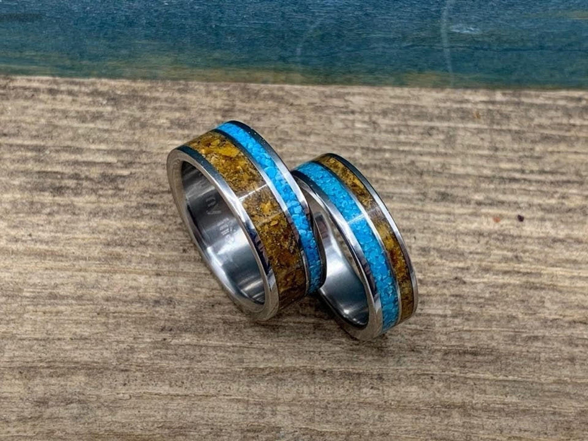 Two metal rings featuring inlaid bands of turquoise stone and brown tiger's eye, resting on a textured wooden surface.