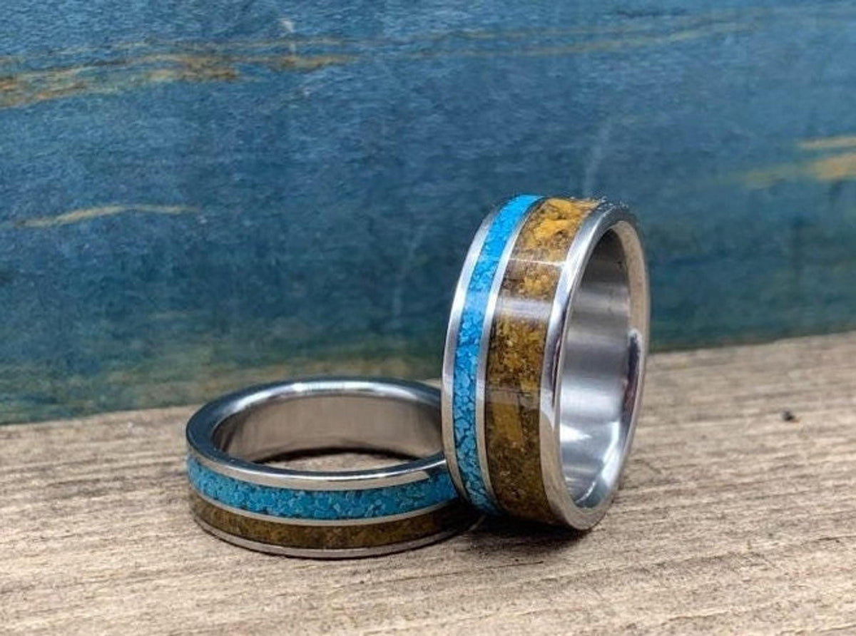 Two metal rings with turquoise and tiger's eye inlays, one standing upright and the other lying flat on a wooden surface.