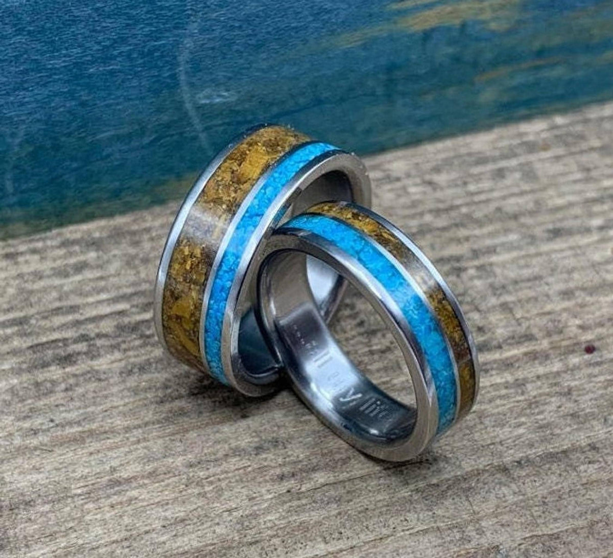 Two metal rings with inlay stripes of bright turquoise and brown tiger's eye, angled together on a wooden shelf.