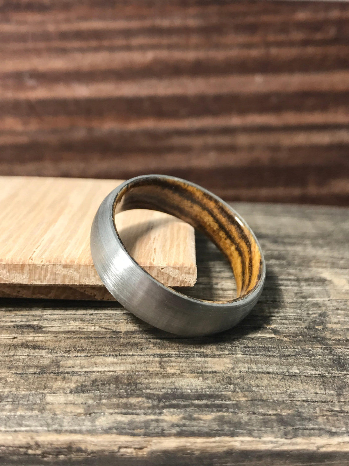 A metal ring with a wood grain interior rests upright on a light wooden block against a rustic wooden background.